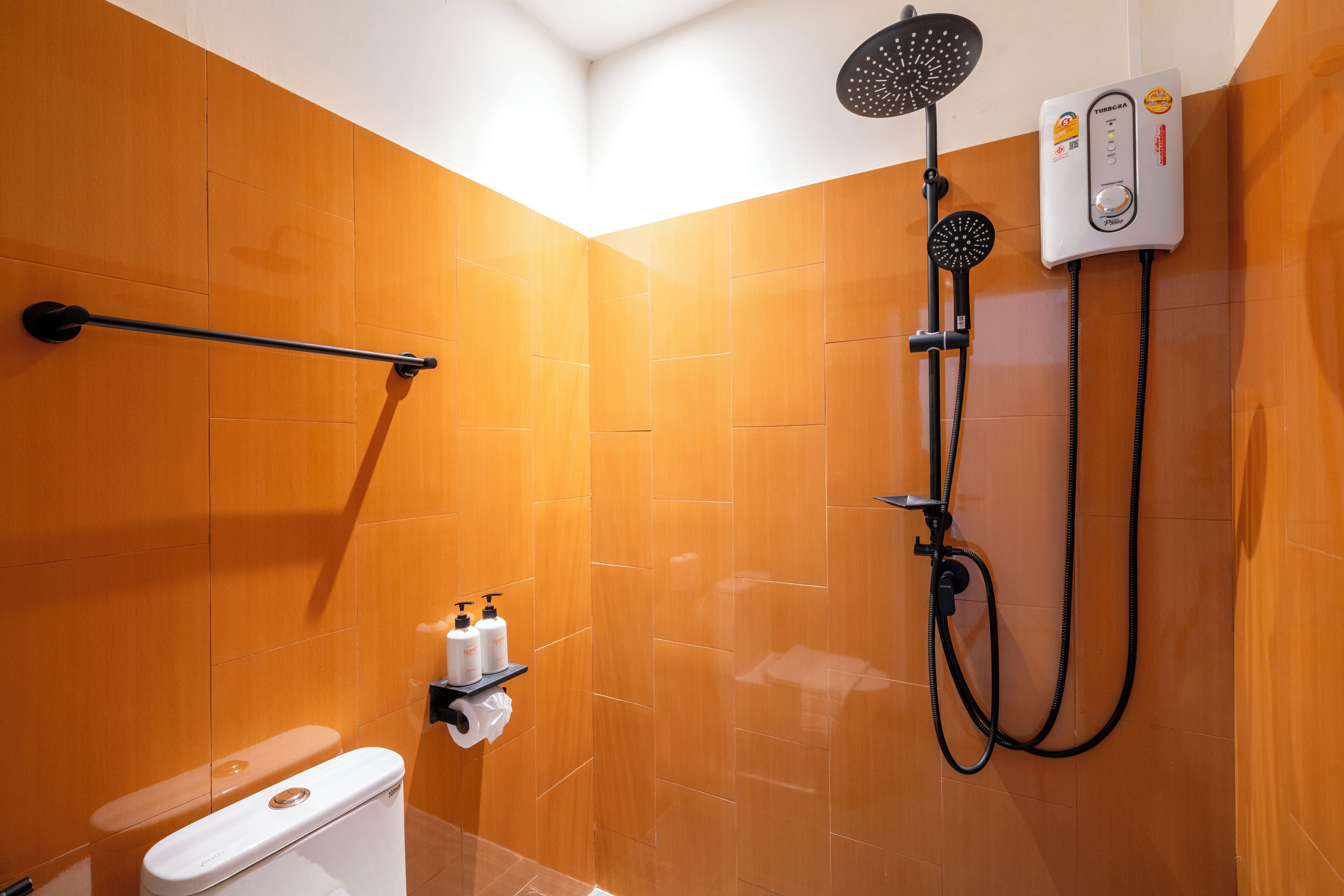Un baño con azulejos naranja, un inodoro blanco, un cabezal de ducha manual y un calentador de agua instalado en la pared.