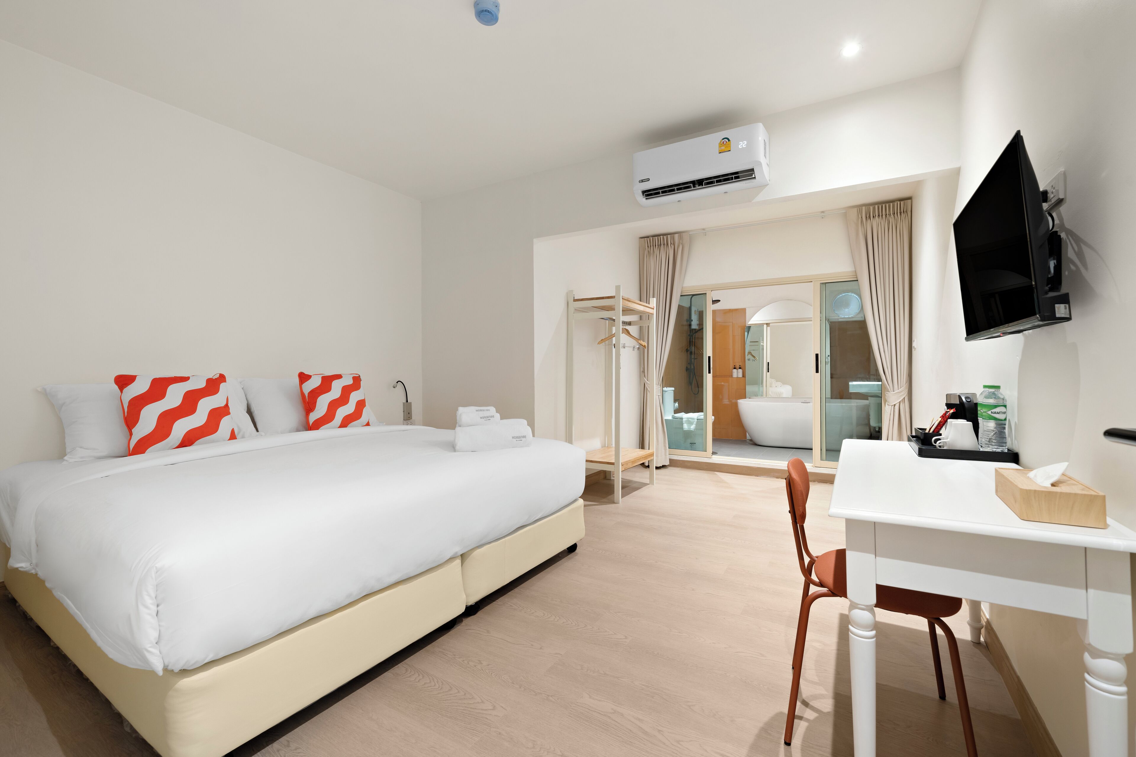Habitación de hotel con cama, escritorio, silla, unidad de aire acondicionado, televisor y baño visible a través de una puerta de vidrio.