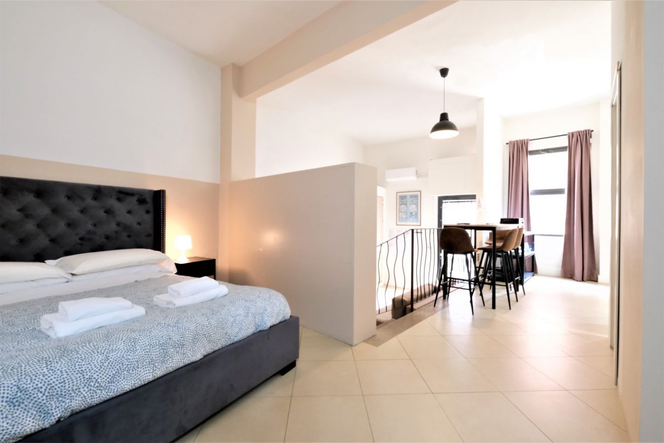 A modern hotel room with a large bed, a dining area with chairs and a table, and a balcony with curtains.