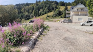 Une allée de gravier mène à un bâtiment à deux étages doté d’un balcon, qui est entouré d’une colline boisée.