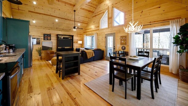 Un intérieur de chalet chaleureux comprenant un coin repas, une cuisine et un salon, avec un plancher en bois et un plafond haut.