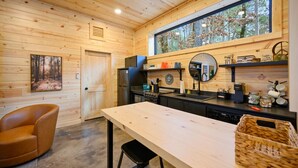 Een houten interieur met een eettafel, een stoel, een keukenhoek met een fornuis, en een groot raam met uitzicht op een bos.
