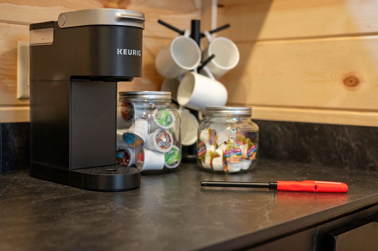 En Keurig-kaffemaskine på en køkkenbordplade med kaffekapsler i glas og en rød skruetrækker.