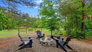 A fire pit with four Adirondack chairs, surrounded by gravel and trees.