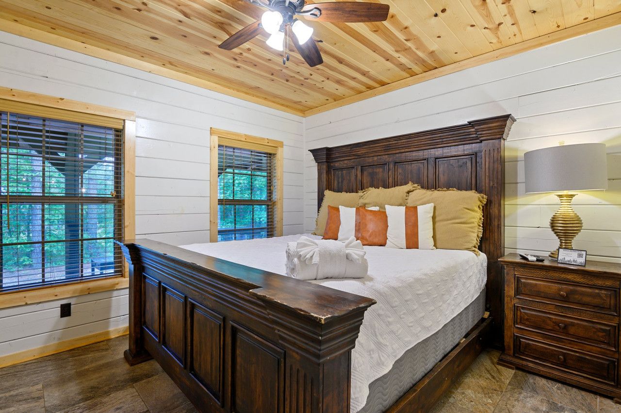 A bedroom with a large bed, wooden headboard, and a ceiling fan. There are two windows with blinds, and a nightstand with a lamp.