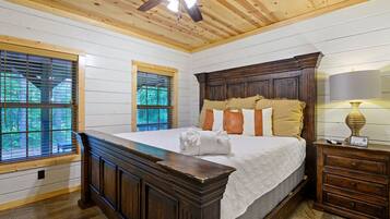 A bedroom with a large bed, wooden headboard, and a ceiling fan. There are two windows with blinds, and a nightstand with a lamp.