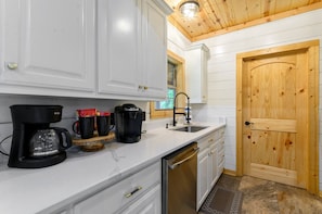 A kitchen with white cabinets, a wooden door, a coffee maker, and a sink. - Hot Tub + Fire Pit Cabin | Pet-Friendly Hochatown Retreat Sleeps 8 | Near Beavers Bend & Broken Bow Lake (Hochatown)