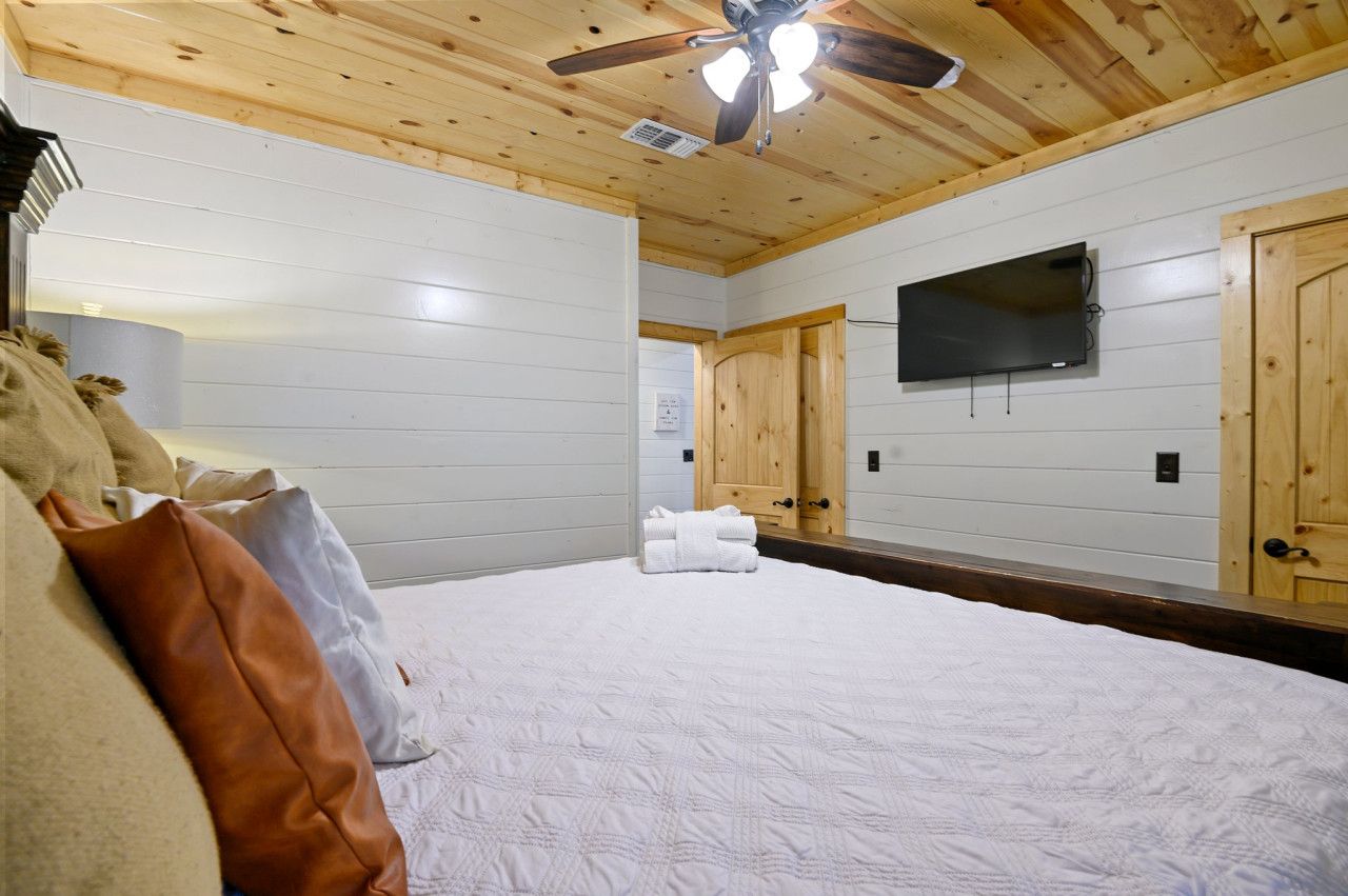 A bedroom with a bed, a ceiling fan, a mounted TV, and wooden doors.