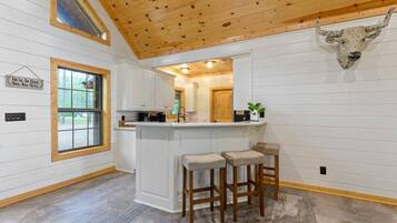 A kitchen with white cabinets, a wooden ceiling, and a bull head mounted on the wall. There are two stools and a window with a sign above it.