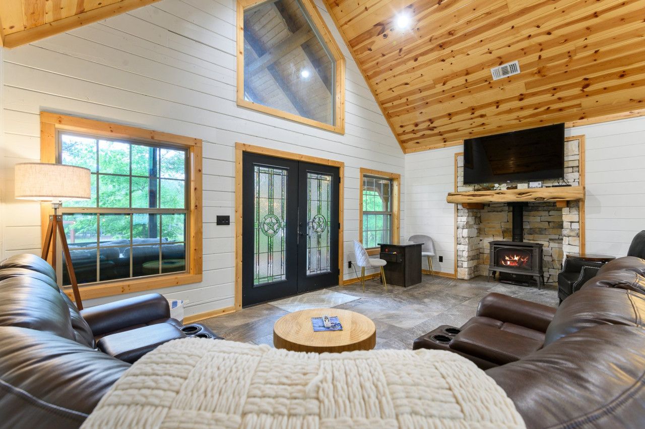 A cozy cabin interior with a stone fireplace, leather sofas, and large windows.