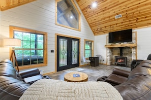A cozy cabin interior with a stone fireplace, leather sofas, and large windows.