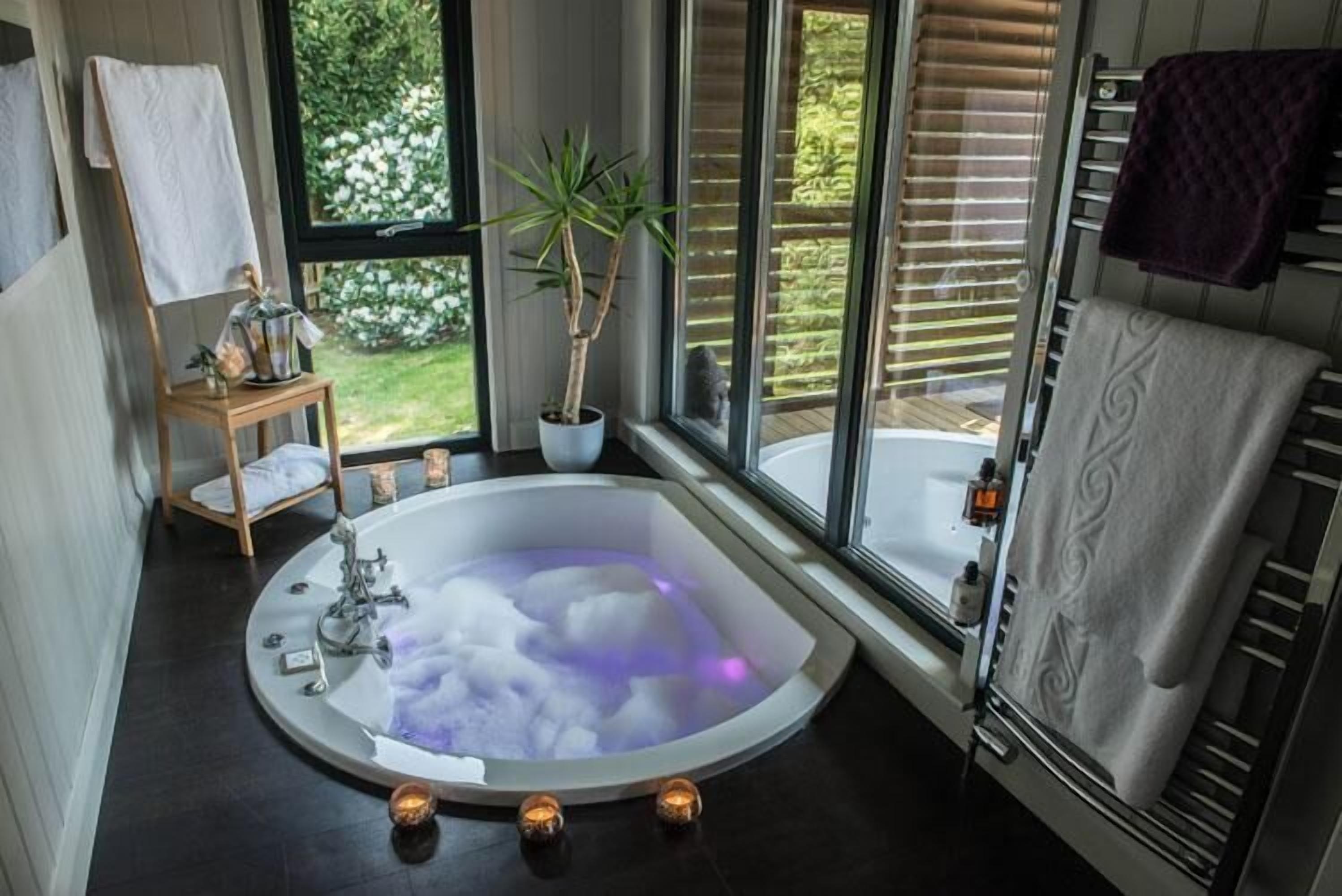 Une salle de bain moderne avec une baignoire sur pieds, une petite table en bois avec une serviette, une plante en pot et une vue sur la verdure à l’extérieur.