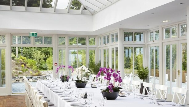 Un invernadero con una larga mesa de comedor dispuesta para un evento formal, rodeado de sillas blancas y plantas en macetas.