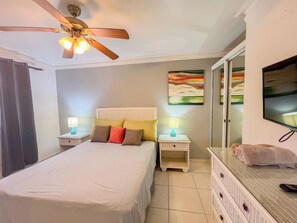 Un dormitorio con cama, mesitas de noche, ventilador de techo, televisor, un cuadro y cómoda.