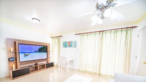 Una sala de estar con televisión de pantalla plana, muebles blancos, ventilador de techo y vista al mar.