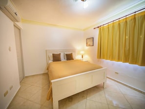A bedroom with a bed, a nightstand, a lamp, a window with curtains, and an air conditioning unit.
