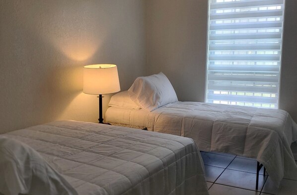 Una habitación con dos camas, una lámpara y una ventana con persianas.