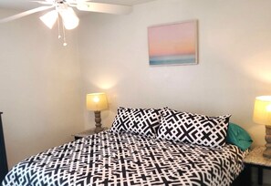 A bedroom with a patterned bedspread, two bedside lamps, a ceiling fan, and a wall-mounted painting.