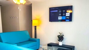 Una sala de estar moderna con un sofá azul, una mesa de centro de vidrio, una lámpara de pie y un televisor montado mostrando servicios de transmisión.
