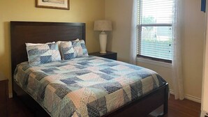 A bedroom with a bed, a nightstand, a lamp, a window with blinds, and a framed picture on the wall. - This house is a 2 bedroom(s), 1 bathrooms, located in Rockport, TX. (Rockport)