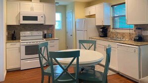 A compact kitchen with white cabinets, a microwave, stove, refrigerator, and a coffee maker. There is a round table with chairs and a window with blinds. - This house is a 2 bedroom(s), 1 bathrooms, located in Rockport, TX. (Rockport)