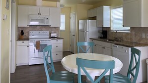 Cocina pequeña con gabinetes blancos, microondas, estufa y refrigerador. Hay una mesa de comedor con sillas y una ventana con persianas.