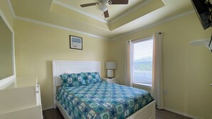 Un dormitorio con cama, ventilador de techo, ventana con persianas, mesita de noche y televisor montado en la pared.
