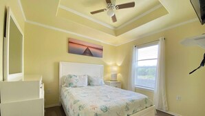 Een slaapkamer met een bed, een plafondventilator, een nachtkastje met lamp, een raam met gordijnen en een schilderij aan de muur.