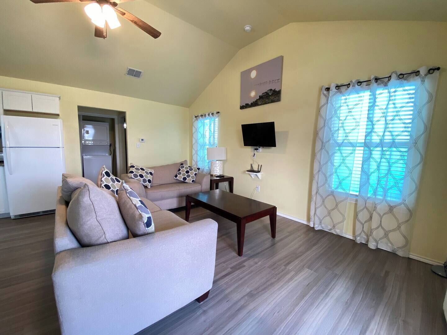 Ein Wohnzimmer mit einer Couch, einem Couchtisch und einem Fernseher. Es gibt eine Küchenzeile mit einem Kühlschrank und einer Mikrowelle.