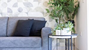 A grey sofa with black cushions, a small round table with a plant and white mugs, and a patterned wall in the background.