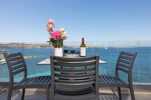 A table with two chairs, a vase of flowers, two wine glasses, and a bottle of wine on a balcony overlooking the sea.