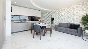 Een moderne keuken met witte kastjes, een eettafel met stoelen, en een grijze bank in een woonruimte met een behang met patroon.