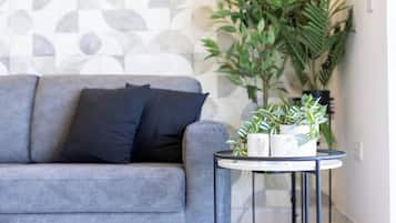 Un sofà gris amb coixins negres, una taula rodona petita amb una planta i tasses blanques, i una paret amb un disseny decoratiu al fons.