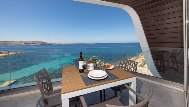 An outdoor dining area with a table set for two, overlooking a clear blue sea and a distant shoreline. - Modern Bugibba apartment right by the beach  Featuring 2 bedrooms, 2 bathrooms, fast Wi-Fi, AC, Smart TV and a fully equipped kitchen — perfect for a relaxed seaside getaway (San Pawl il-Baħar)