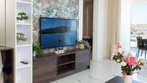 A modern living room with a flat-screen TV mounted on a marble wall, surrounded by shelves with plants, a wooden entertainment unit, and a balcony with a view. - Modern Bugibba apartment right by the beach  Featuring 2 bedrooms, 2 bathrooms, fast Wi-Fi, AC, Smart TV and a fully equipped kitchen — perfect for a relaxed seaside getaway (San Pawl il-Baħar)