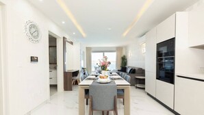 A modern kitchen with white cabinetry, a dining area with a marble floor, and a living space with a sofa and a TV. - Modern Bugibba apartment right by the beach  Featuring 2 bedrooms, 2 bathrooms, fast Wi-Fi, AC, Smart TV and a fully equipped kitchen — perfect for a relaxed seaside getaway (San Pawl il-Baħar)