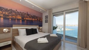 A hotel room with a large bed, a nightstand, a wall-mounted lamp, a window with a view of the sea and boats, and a cityscape mural. - Modern Bugibba apartment right by the beach  Featuring 2 bedrooms, 2 bathrooms, fast Wi-Fi, AC, Smart TV and a fully equipped kitchen — perfect for a relaxed seaside getaway (San Pawl il-Baħar)