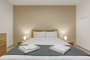 Un lit bien fait, avec deux serviettes pliées, une table de chevet en bois avec une lampe, et un mur sobre.