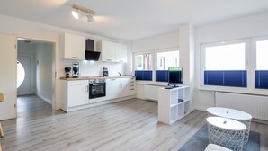 Ett modernt kök med vita skåp, en svart ugn och en inbyggd köksfläkt. Det finns två fönster med blå persienner, en vit matplats med ett runt bord och en grå matta.