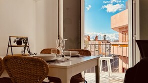 Ein Essbereich mit Tisch für zwei Personen, Korbstühle, einer weißen Tischdecke und Blick auf eine Stadtlandschaft vom Balkon aus.