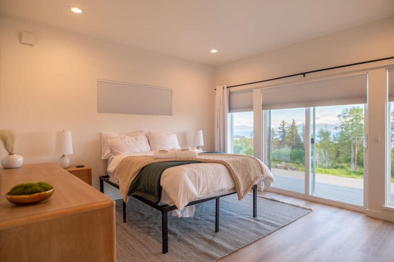 A modern bedroom with a large bed, a desk, and a view of the outdoors through a sliding glass door.