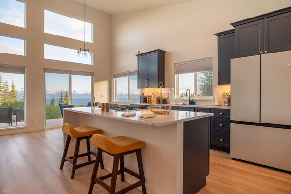 Une cuisine moderne dotée d’un grand îlot central, de meubles de couleur sombre et offrant une vue sur les montagnes.