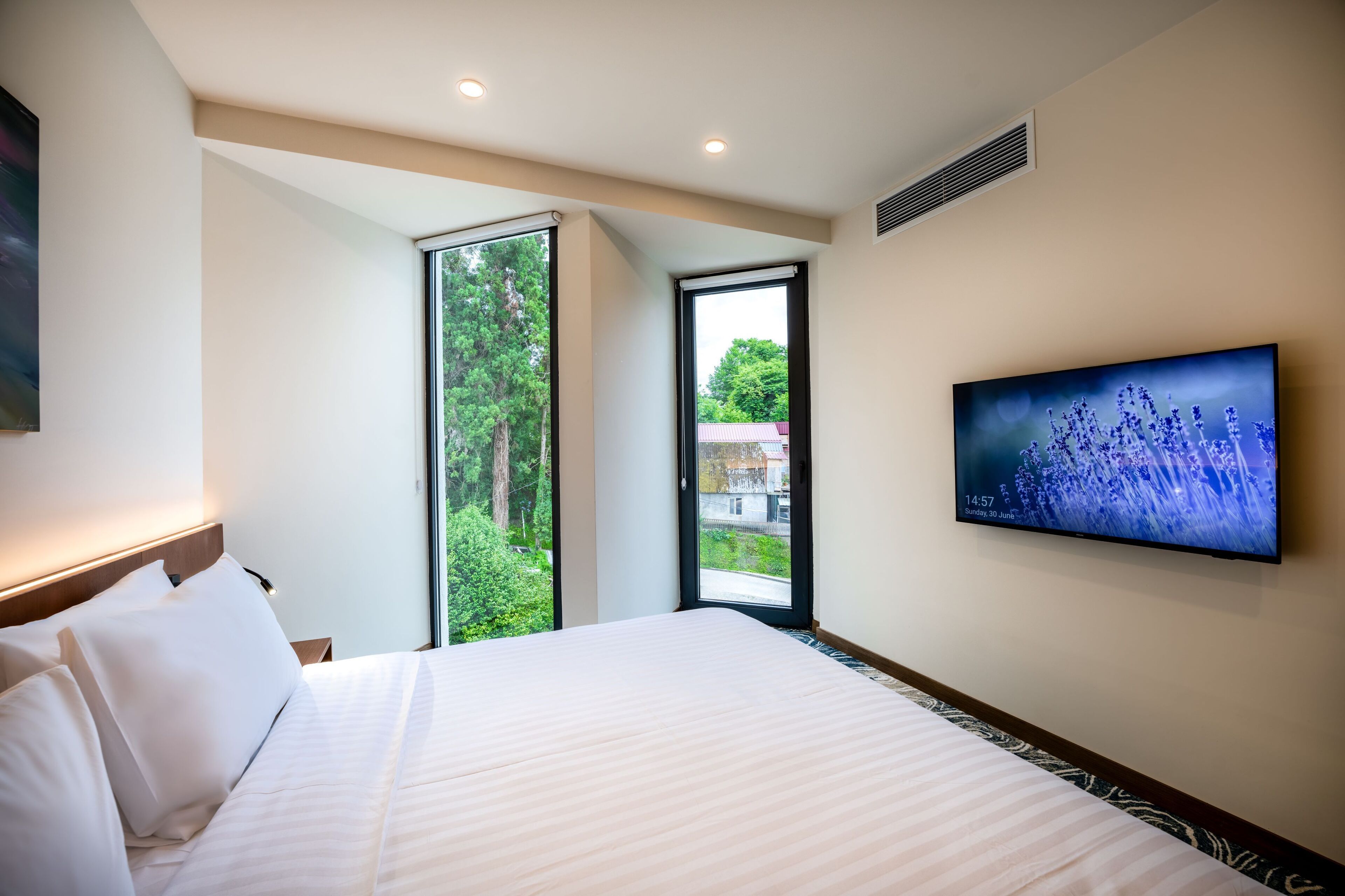 A modern hotel room with a large bed, a television on the wall, and a window offering a view of greenery and buildings outside.