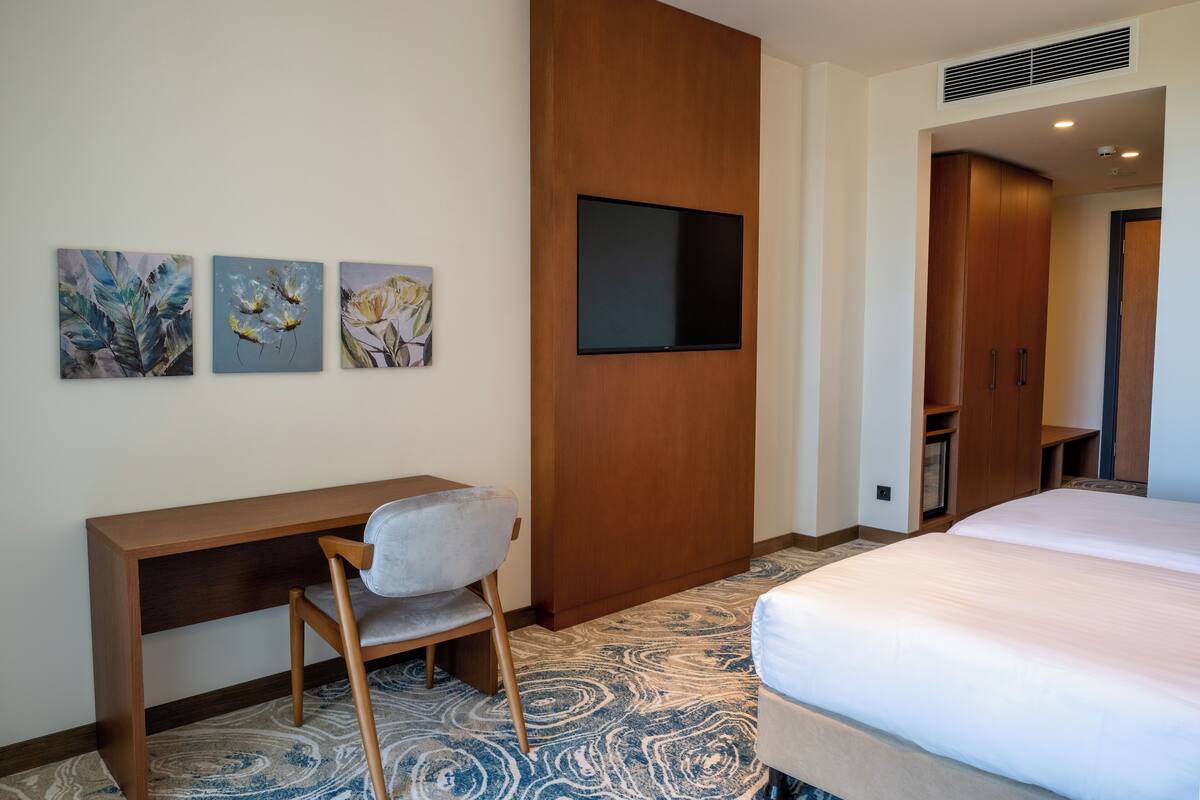 A hotel room with a bed, a desk with a chair, a TV mounted on the wall, and three paintings on the wall.