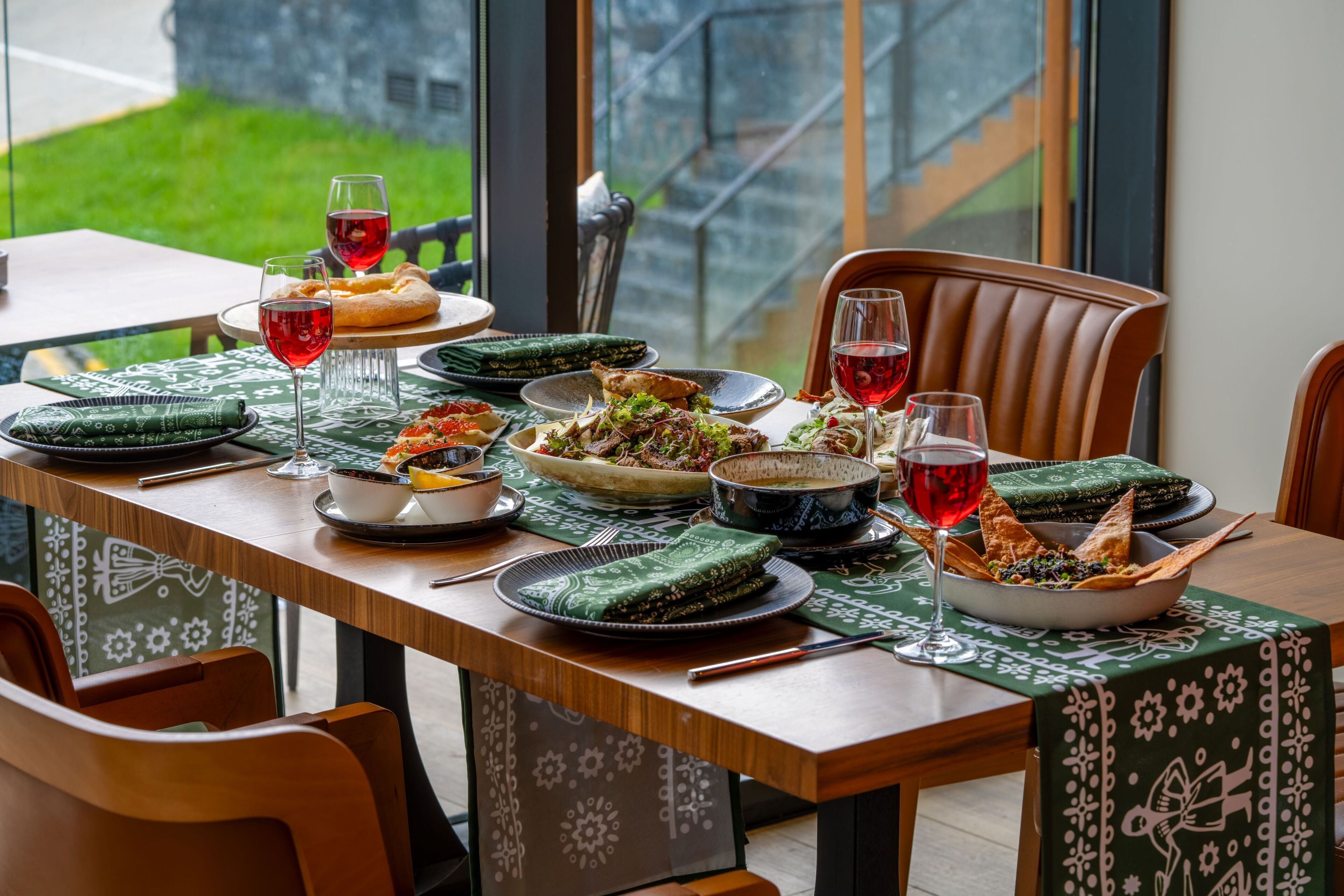 A dining table set with various dishes, glasses of red wine, and a green patterned tablecloth.