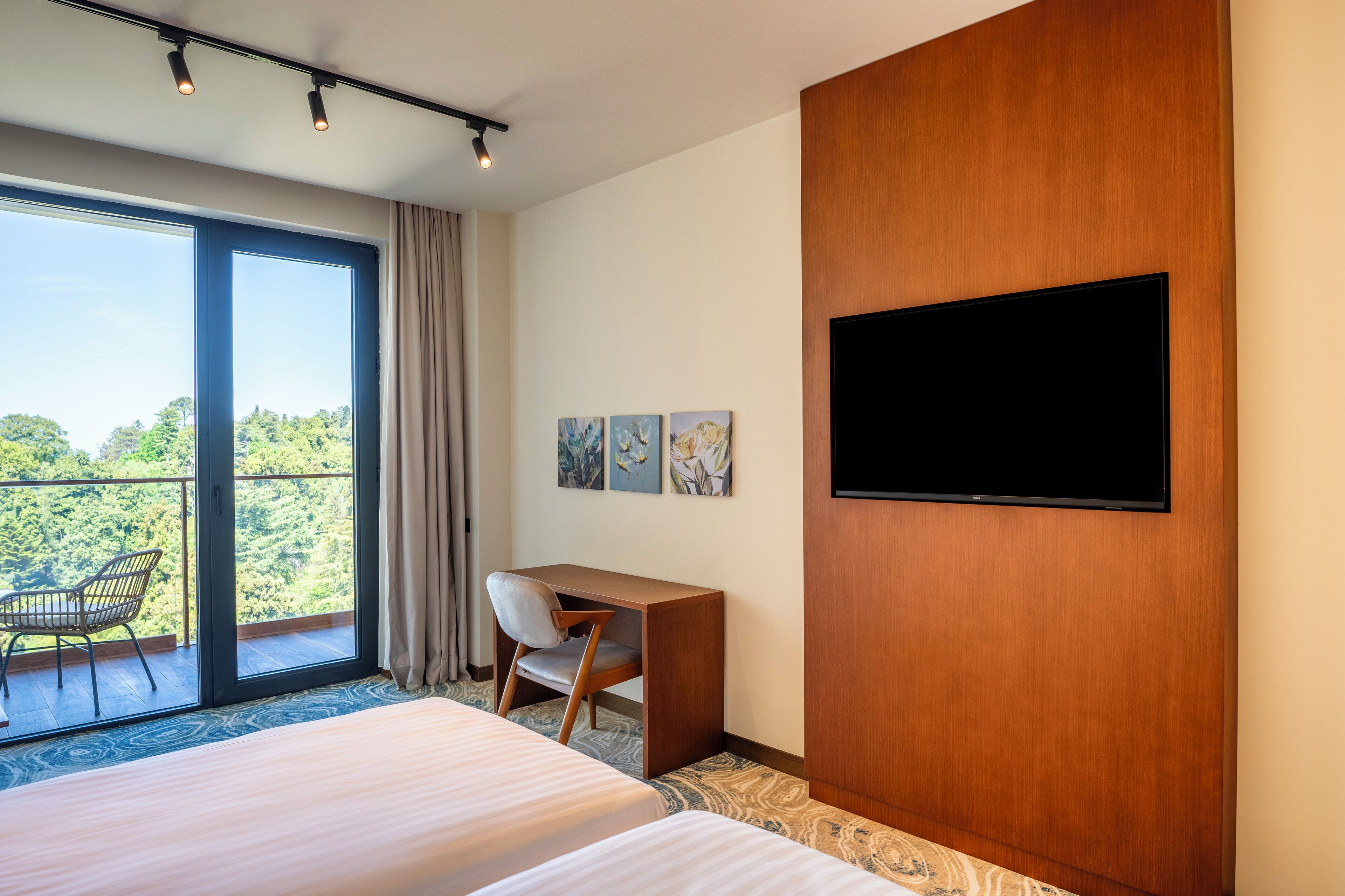 Habitación de hotel con dos camas, un escritorio, una silla, un televisor y un balcón con vistas a árboles.