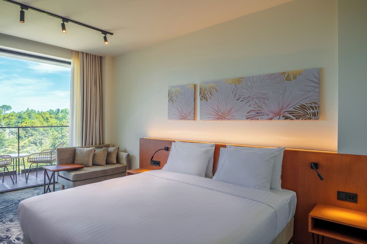 A hotel room with a large bed, a sofa, a small table, and a chair. There is a window with a view of greenery.