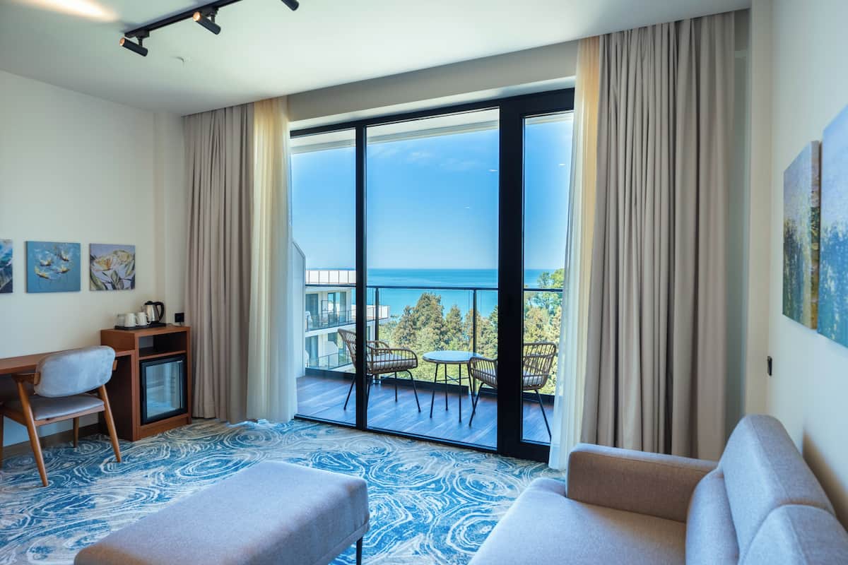 A modern hotel room with a balcony, a desk, a sofa, and a patterned carpet.