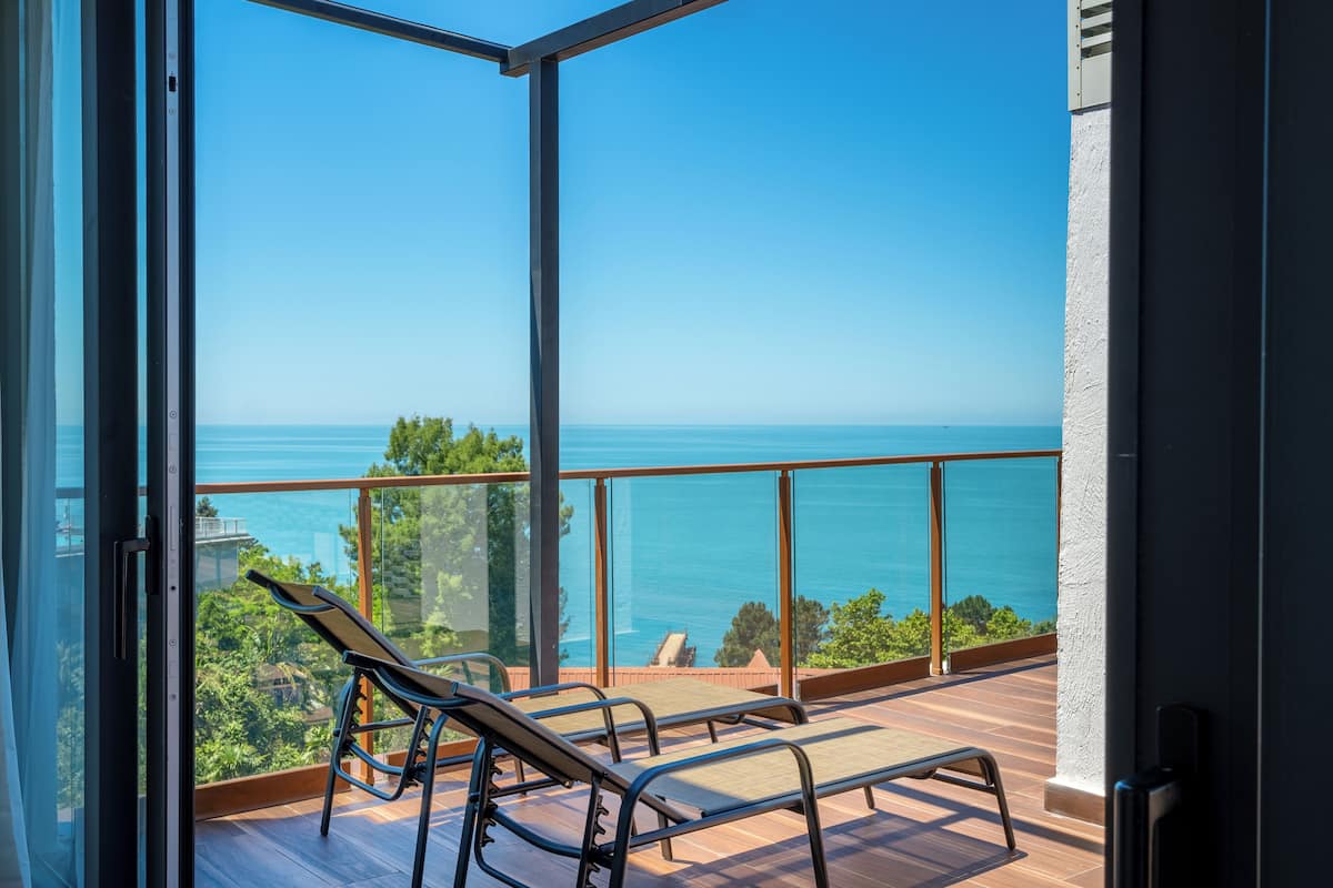 A balcony with two lounge chairs overlooking a clear blue sea and greenery.