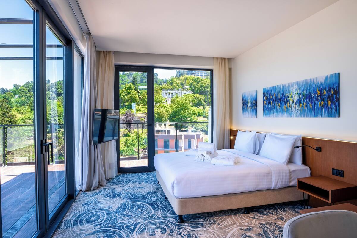 A hotel room with a large bed, a flat-screen TV, a balcony with a view of greenery and buildings, and two abstract paintings on the wall.